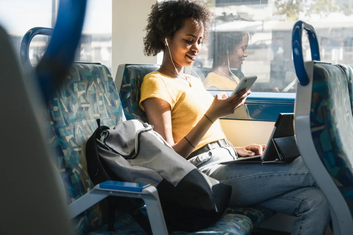 A woman wearing earphones looks at her phone on a bus, with a tablet and keyboard on her lap.