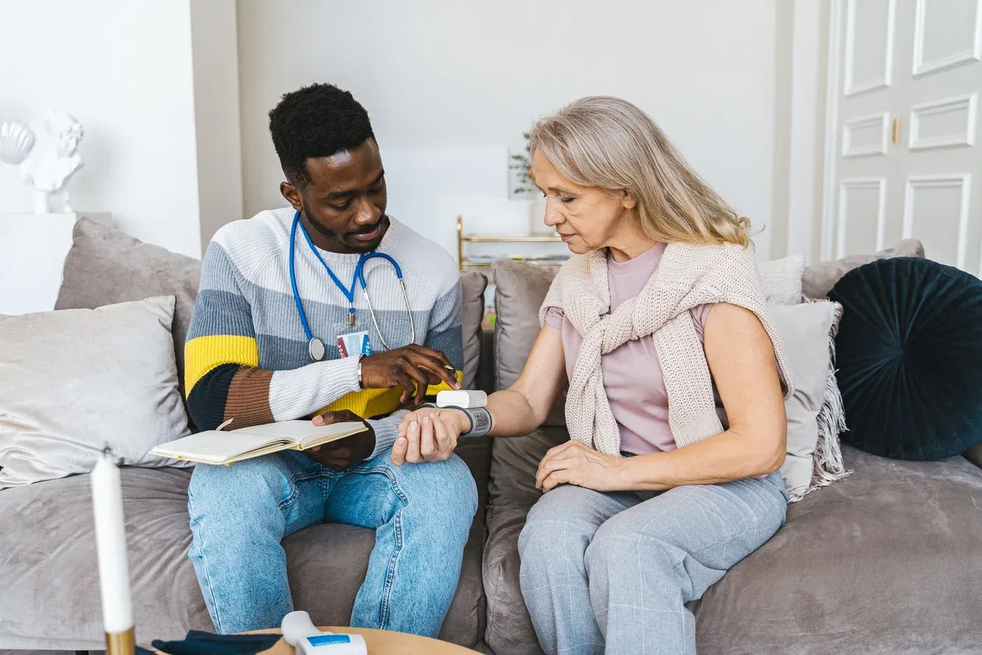A male healthcare worker checks an older woman’s blood pressure at home, with a stethoscope and notebook.