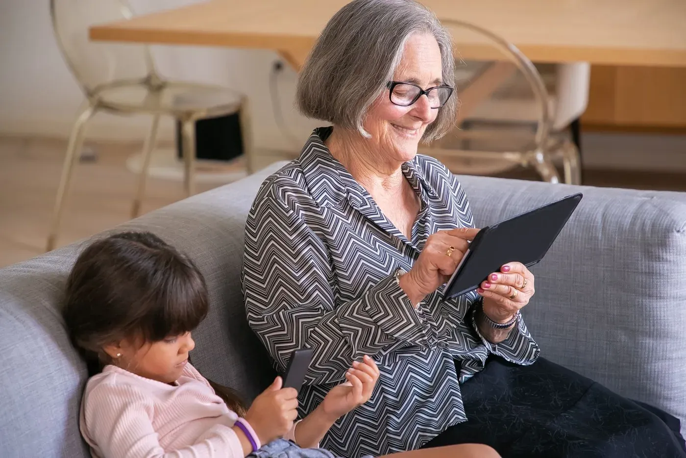 An older woman uses a tablet on a sofa, with a young girl seated beside her.